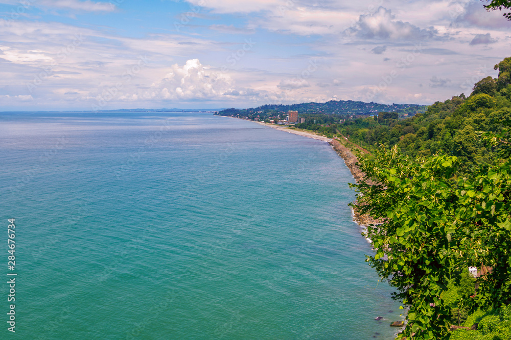Aerial summer landscape – sea with turquoise water, green trees, blue sky with clouds, mountains and small town on the horizon. View of Black Sea in Batumi, Georgia.