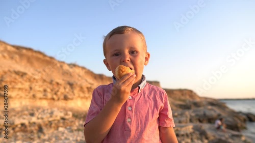 Closeup, child boy eating a bun on a shore. Real positive emotions, real time video footage, boy on a sea coast background, 4K