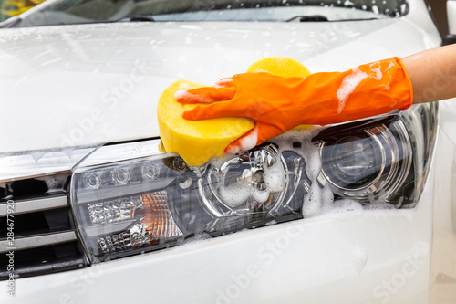 Woman hand wearing orange gloves with yellow sponge washing headlight modern car or cleaning automobile. Car wash concept
