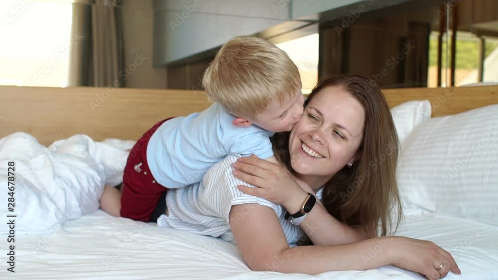 Mom and son lie on the bed in the morning on the weekend and play, the boy kisses and hugs his beloved mother. Slow motion. Mom and son lie on the bed in the morning on the weekend and play, the boy kisses and hugs his beloved mother. Slow motion.