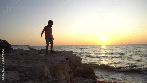 Silhouette of a little boy jumping over coastal rocks against a background of sunset on the sea. Beautiful dark waves lit by the rays of the sun
