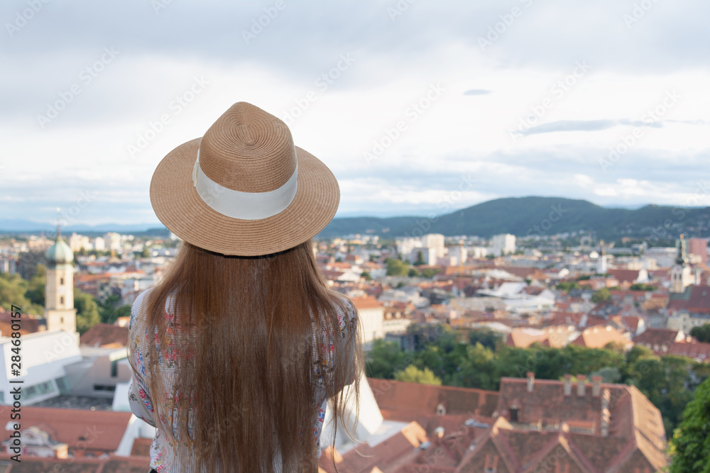 A girl with long hair in a hat stands on the observation deck and looks at the city of Graz.