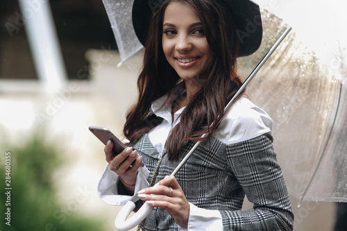 Close up portrait of beautiful smiling woman wearing black hat and gray coat under transparent umbrella, typing sms