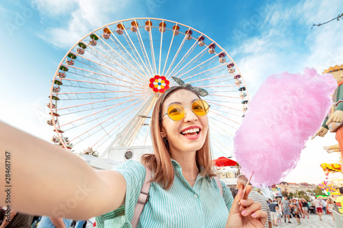 Happy woman eating tasty colorful pink cotton candy in front of the big white ferris wheel at the amusement park in Europe photo