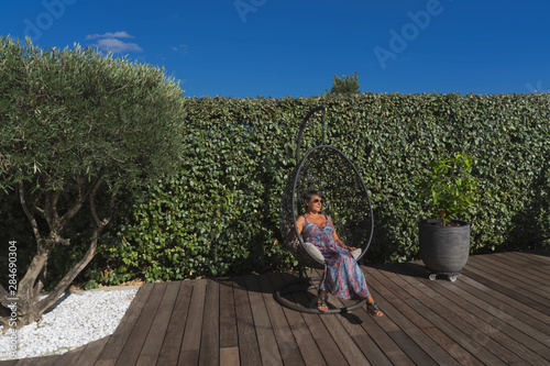 a woman sit in a suspended seat on a wooden floor in a garden with an olive tree