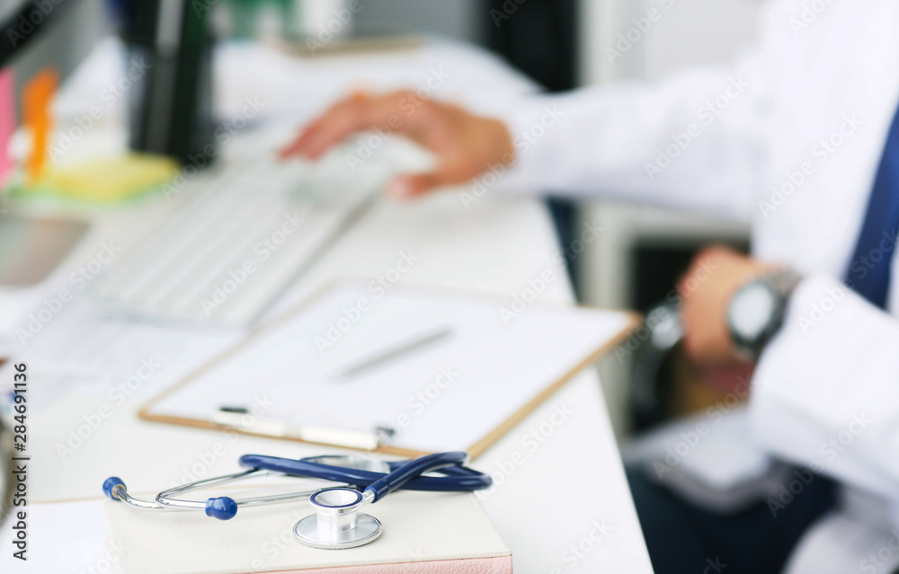 Male doctor working with laptop computer. Clipboard, pen and stethoscope on desk in hospital close-up. Male doctor working with computer in the background. Male doctor working with laptop computer. Clipboard, pen and stethoscope on desk in hospital close-up. Male doctor working with computer in the background.