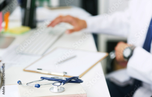 Male doctor working with laptop computer. Clipboard, pen and stethoscope on desk in hospital close-up. Male doctor working with computer in the background.