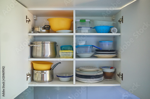 opened white kitchen cupboard with plates, metal pots and food containers