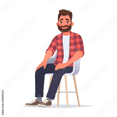 Bearded smiling man dressed in casual clothes is sitting on a chair on a white background