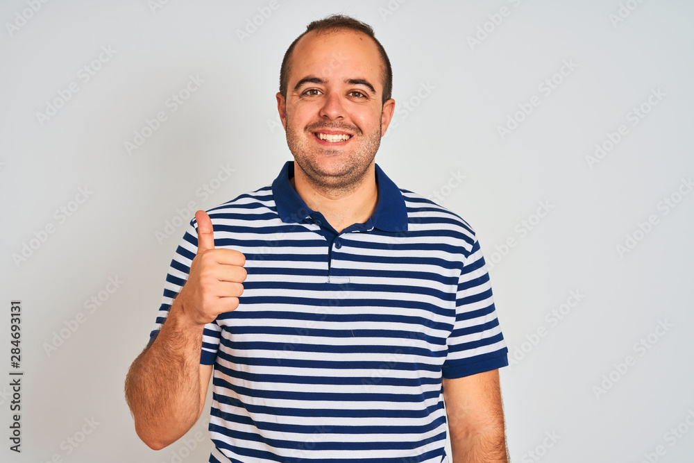 Young man wearing casual striped polo standing over isolated white background doing happy thumbs up gesture with hand. Approving expression looking at the camera showing success. Young man wearing casual striped polo standing over isolated white background doing happy thumbs up gesture with hand. Approving expression looking at the camera showing success.