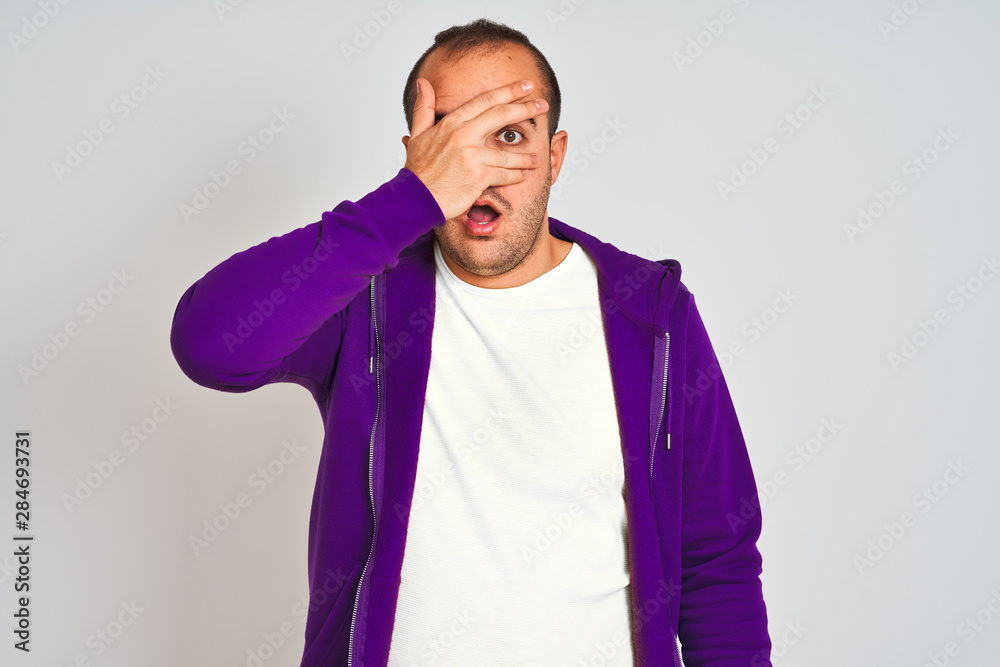 Young man wearing purple sweatshirt standing over isolated white background peeking in shock covering face and eyes with hand, looking through fingers with embarrassed expression. Young man wearing purple sweatshirt standing over isolated white background peeking in shock covering face and eyes with hand, looking through fingers with embarrassed expression.