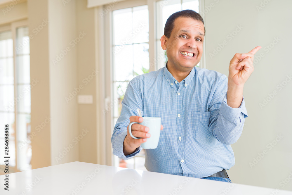 Middle age man drinking coffee in the morning at home very happy pointing with hand and finger to the side Middle age man drinking coffee in the morning at home very happy pointing with hand and finger to the side