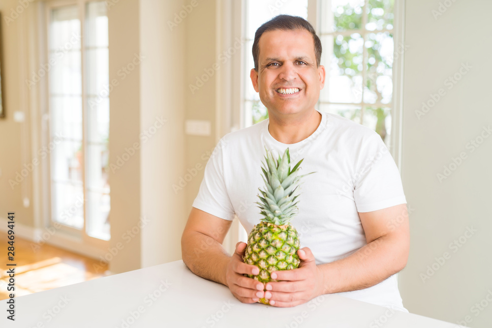 Middle age man eating fresh tropical pineapple at home with a happy face standing and smiling with a confident smile showing teeth Middle age man eating fresh tropical pineapple at home with a happy face standing and smiling with a confident smile showing teeth