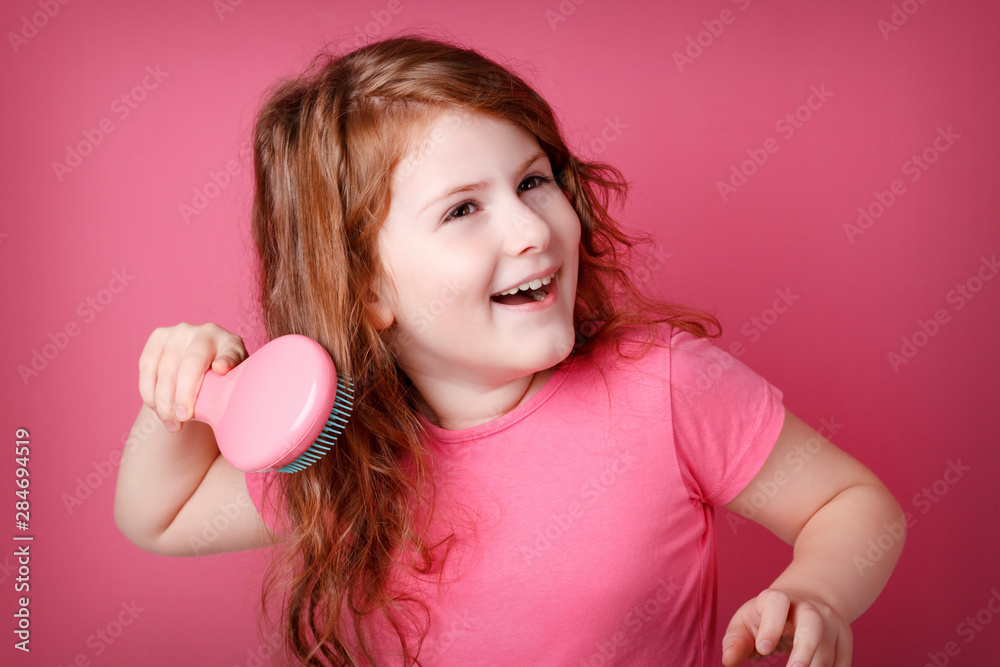 Redhead curly girl is grooming and combing her hair. Little teen girl smiling on pink background. Copy space.
