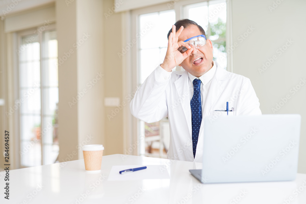 Middle age scientist man wearing professional glasses working using laptop with happy face smiling doing ok sign with hand on eye looking through fingers