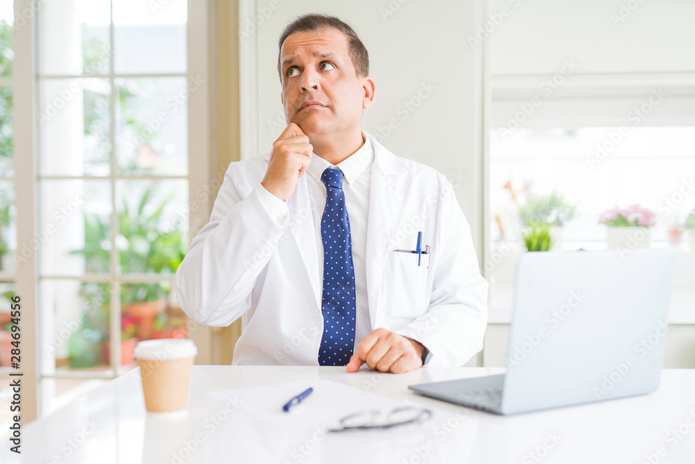 Middle age doctor man wearing white medical coat working with laptop at the clinic with hand on chin thinking about question, pensive expression. Smiling with thoughtful face. Doubt concept. Middle age doctor man wearing white medical coat working with laptop at the clinic with hand on chin thinking about question, pensive expression. Smiling with thoughtful face. Doubt concept.