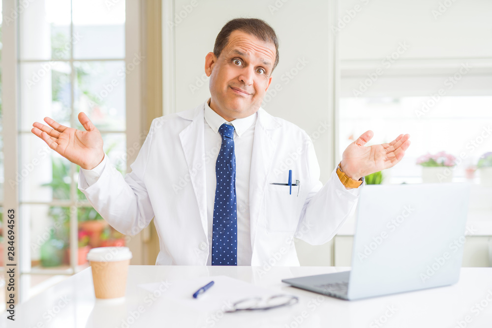 Middle age doctor man wearing white medical coat working with laptop at the clinic clueless and confused expression with arms and hands raised. Doubt concept. Middle age doctor man wearing white medical coat working with laptop at the clinic clueless and confused expression with arms and hands raised. Doubt concept.