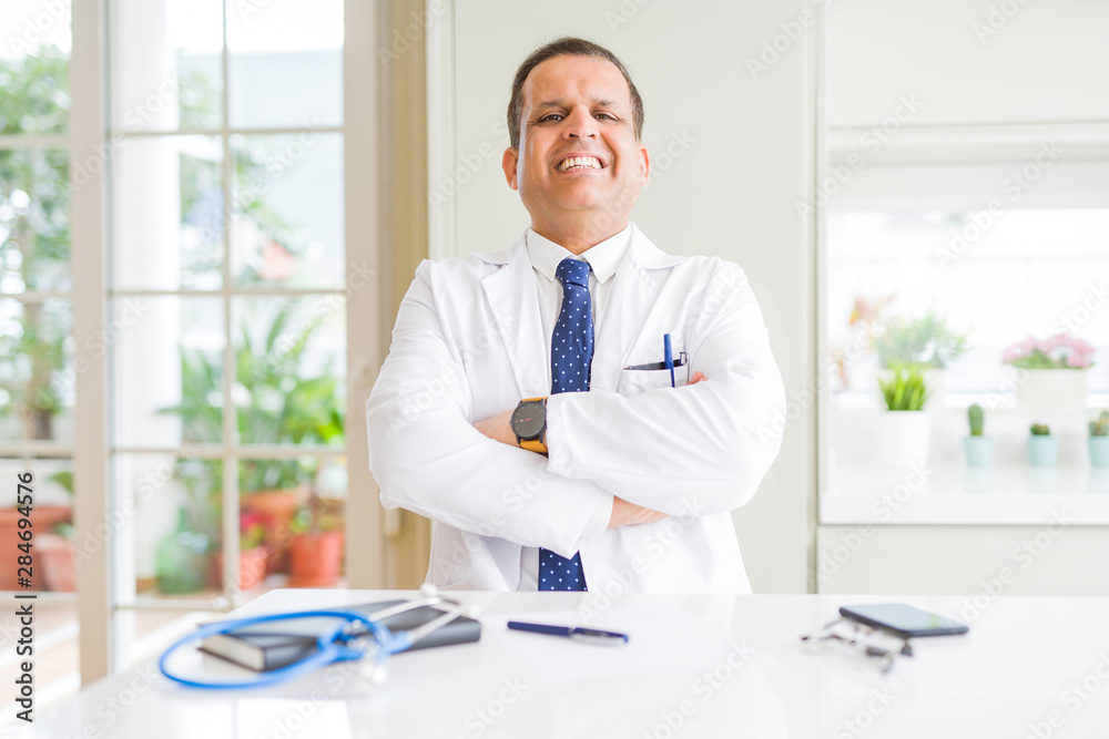 Middle age doctor man wearing medical coat at the clinic happy face smiling with crossed arms looking at the camera. Positive person.