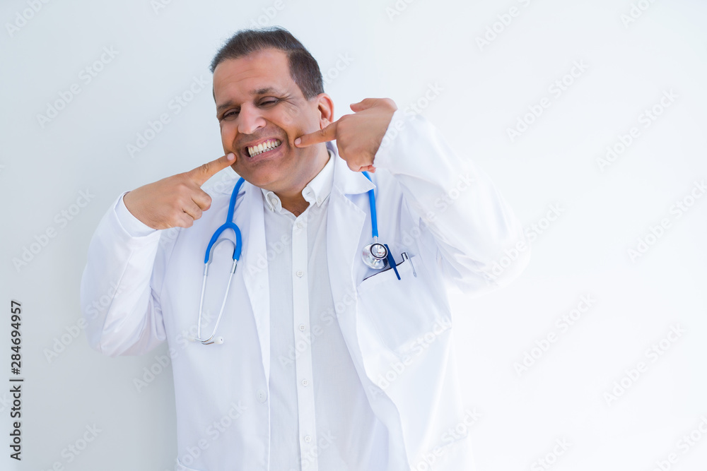 Middle age doctor man wearing stethoscope and medical coat over white background smiling confident showing and pointing with fingers teeth and mouth. Health concept. Middle age doctor man wearing stethoscope and medical coat over white background smiling confident showing and pointing with fingers teeth and mouth. Health concept.