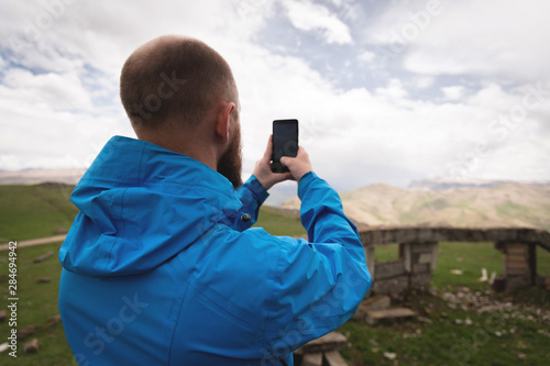 A bearded man takes photos or videos outdoors on his smartphone. A guy in a jacket on a background of mountain valleys and a gray sky weedt broadcast on the phone. Back view