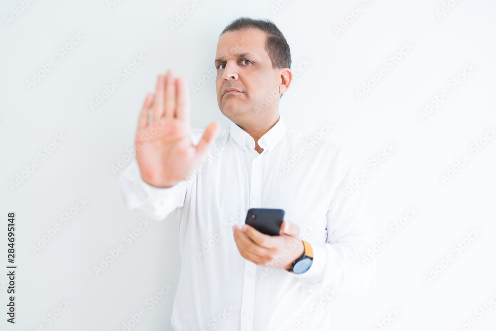 Middle age man using smartphone over white wall with open hand doing stop sign with serious and confident expression, defense gesture Middle age man using smartphone over white wall with open hand doing stop sign with serious and confident expression, defense gesture