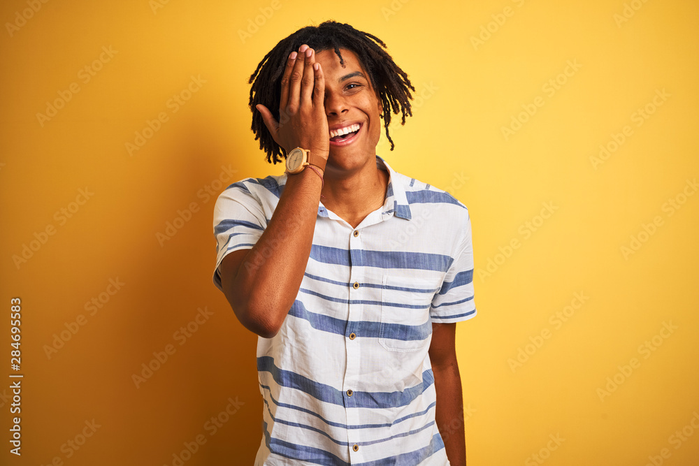 Afro american man with dreadlocks wearing striped shirt over isolated yellow background covering one eye with hand, confident smile on face and surprise emotion. Afro american man with dreadlocks wearing striped shirt over isolated yellow background covering one eye with hand, confident smile on face and surprise emotion.