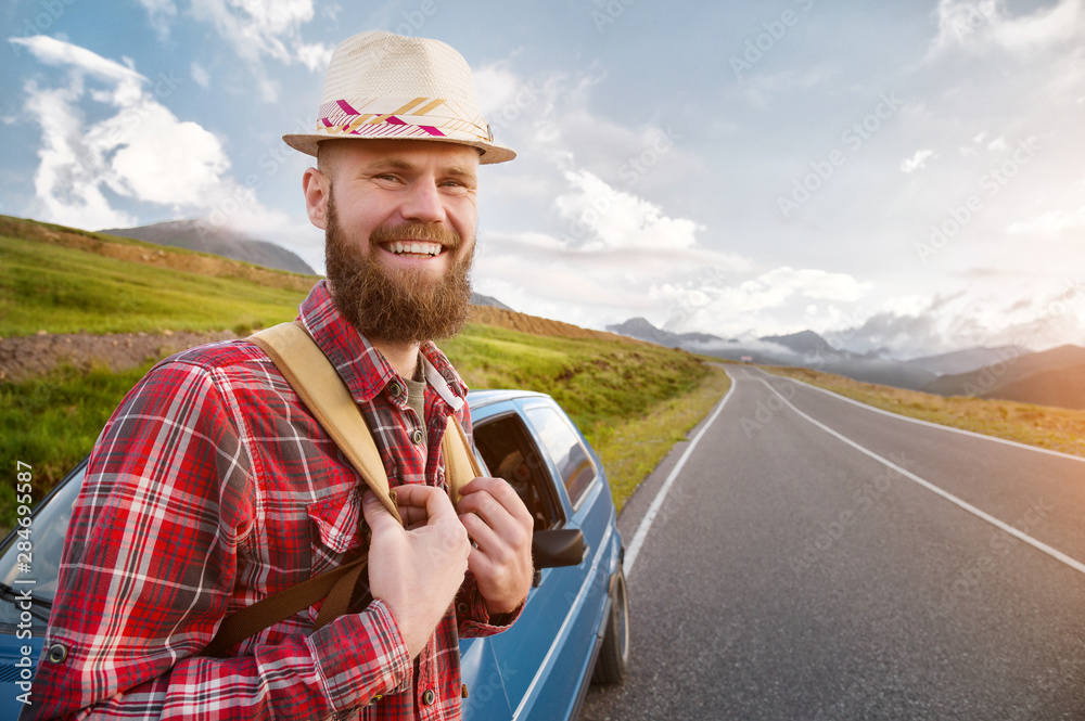 Fototapeta premium Portrait of a bearded happy smiling traveler hipster with a backpack in a plaid shirt and a hat next to an unknown car stands on the road at sunset in the mountains. Happy and confident travel concept