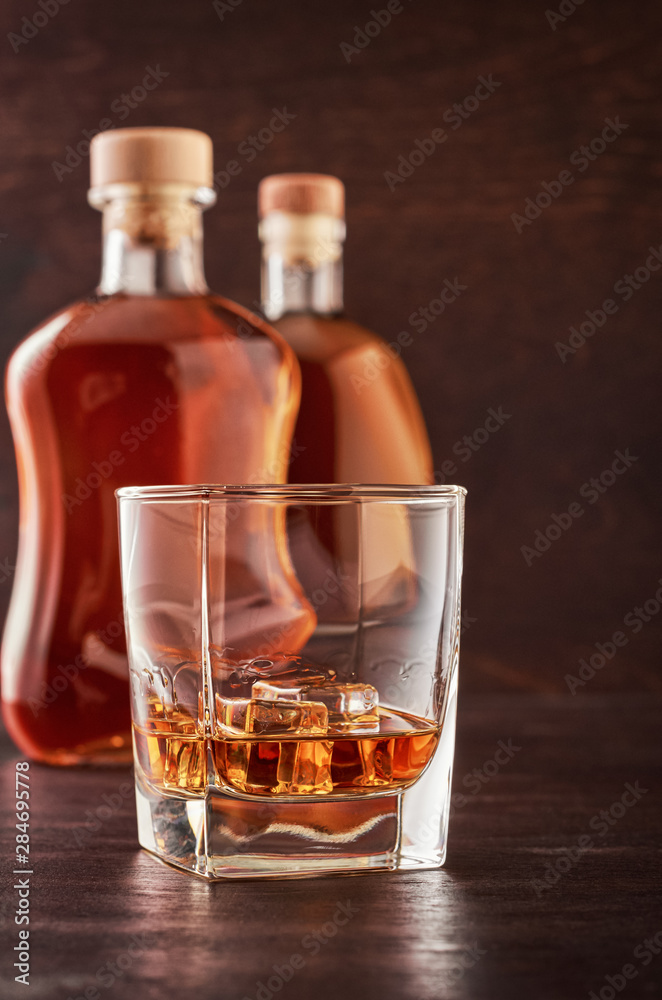 A glass of whisky with ice on a wooden table. In the background, two bottles of whiskey of different shapes. A glass of whisky with ice on a wooden table. In the background, two bottles of whiskey of different shapes.