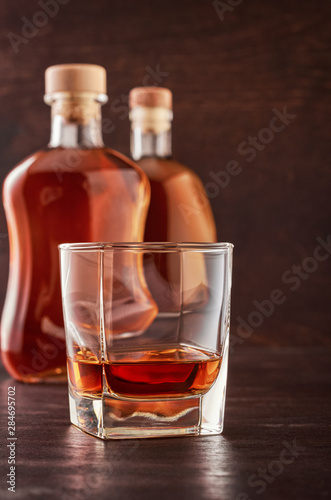 A glass of whiskey on a wooden table. In the background, two bottles of whiskey of different shapes.