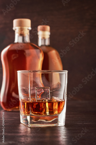 A glass of whisky with ice on a wooden table. In the background, two bottles of whiskey of different shapes.