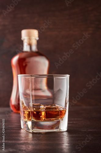 A glass of whisky with ice on a wooden table. There's a full bottle of whiskey in the background.