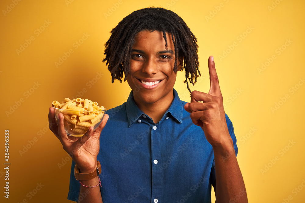 Afro american man with dreadlocks holding pasta macaroni over isolated yellow background surprised with an idea or question pointing finger with happy face, number one Afro american man with dreadlocks holding pasta macaroni over isolated yellow background surprised with an idea or question pointing finger with happy face, number one