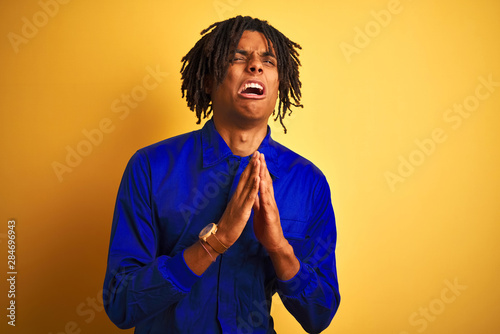 Afro worker man with dreadlocks wearing mechanic uniform over isolated yellow background begging and praying with hands together with hope expression on face very emotional and worried.  © Krakenimages.com