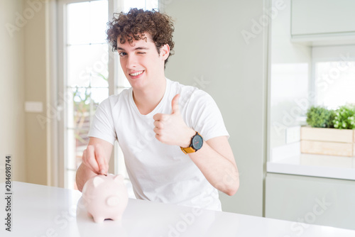 Young man investing money using piggy bank at home happy with big smile doing ok sign, thumb up with fingers, excellent sign