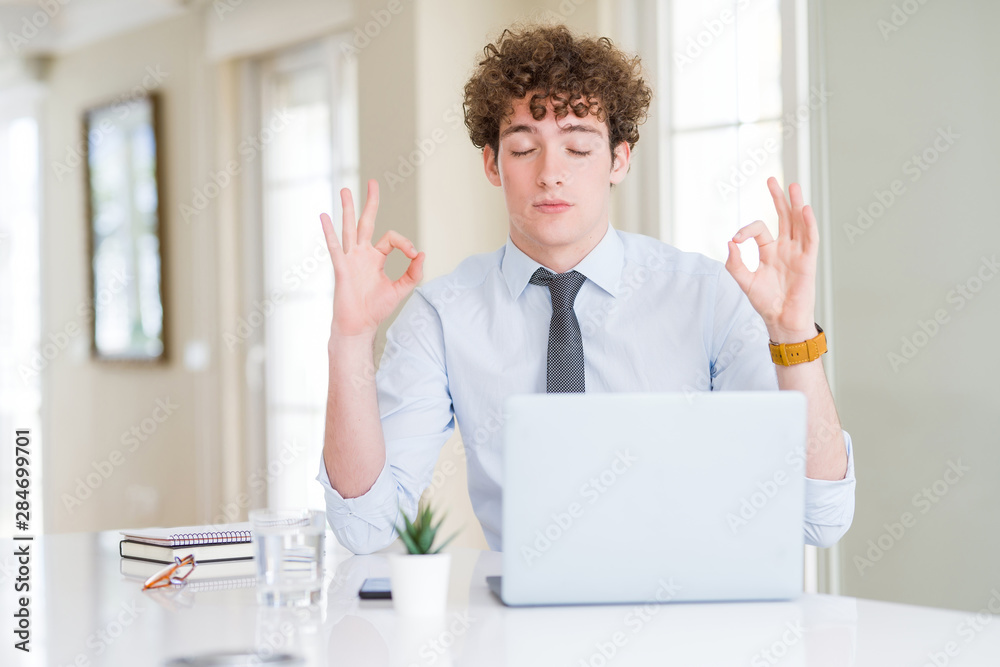 Young business man working with computer laptop at the office relax and smiling with eyes closed doing meditation gesture with fingers. Yoga concept.