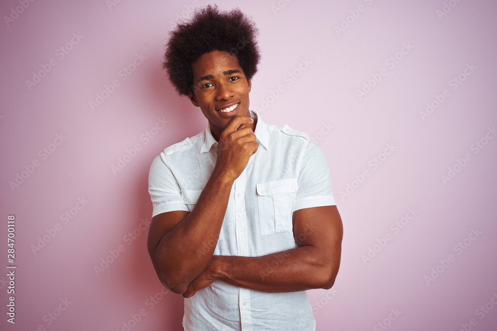 Young american man with afro hair wearing white shirt standing over isolated pink background looking confident at the camera smiling with crossed arms and hand raised on chin. Thinking positive. Young american man with afro hair wearing white shirt standing over isolated pink background looking confident at the camera smiling with crossed arms and hand raised on chin. Thinking positive.