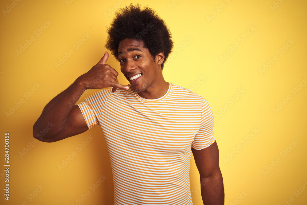 American man with afro hair wearing striped t-shirt standing over isolated yellow background smiling doing phone gesture with hand and fingers like talking on the telephone. Communicating concepts. American man with afro hair wearing striped t-shirt standing over isolated yellow background smiling doing phone gesture with hand and fingers like talking on the telephone. Communicating concepts.