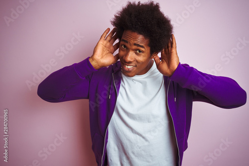 Young african american man wearing purple sweatshirt standing over isolated pink background Trying to hear both hands on ear gesture, curious for gossip. Hearing problem, deaf