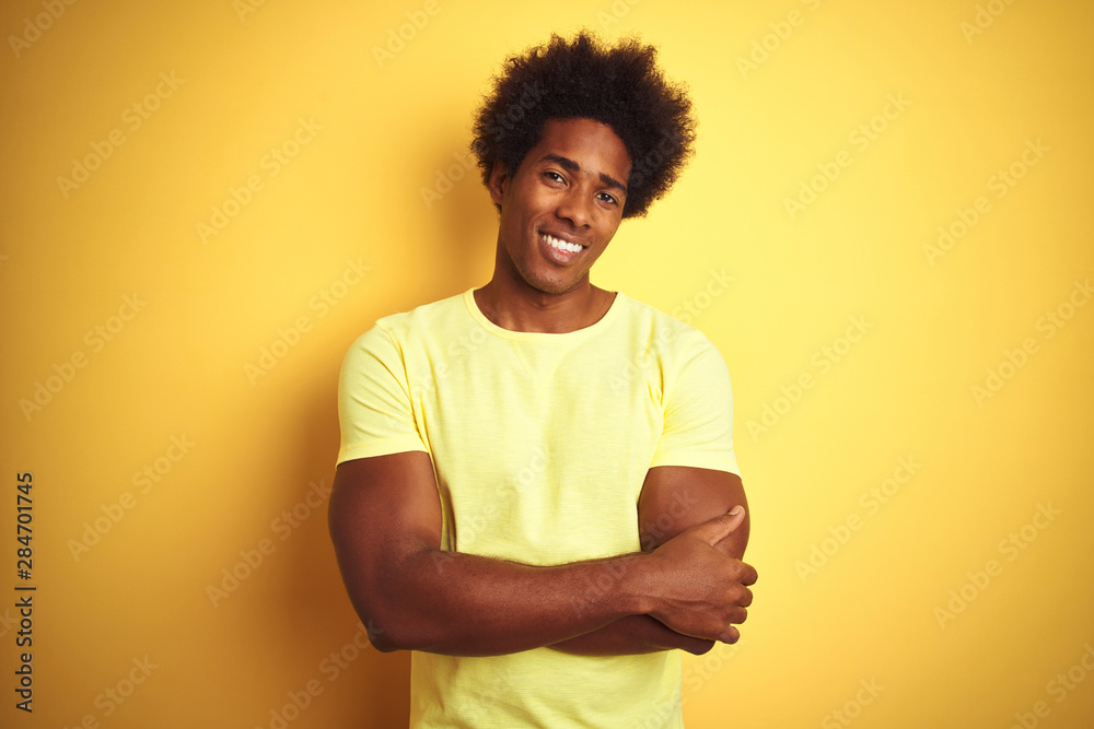 African american man with afro hair wearing t-shirt standing over isolated yellow background happy face smiling with crossed arms looking at the camera. Positive person. African american man with afro hair wearing t-shirt standing over isolated yellow background happy face smiling with crossed arms looking at the camera. Positive person.