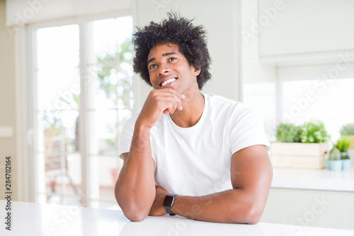 Young african american man wearing casual white t-shirt sitting at home with hand on chin thinking about question, pensive expression. Smiling and thoughtful face. Doubt concept.