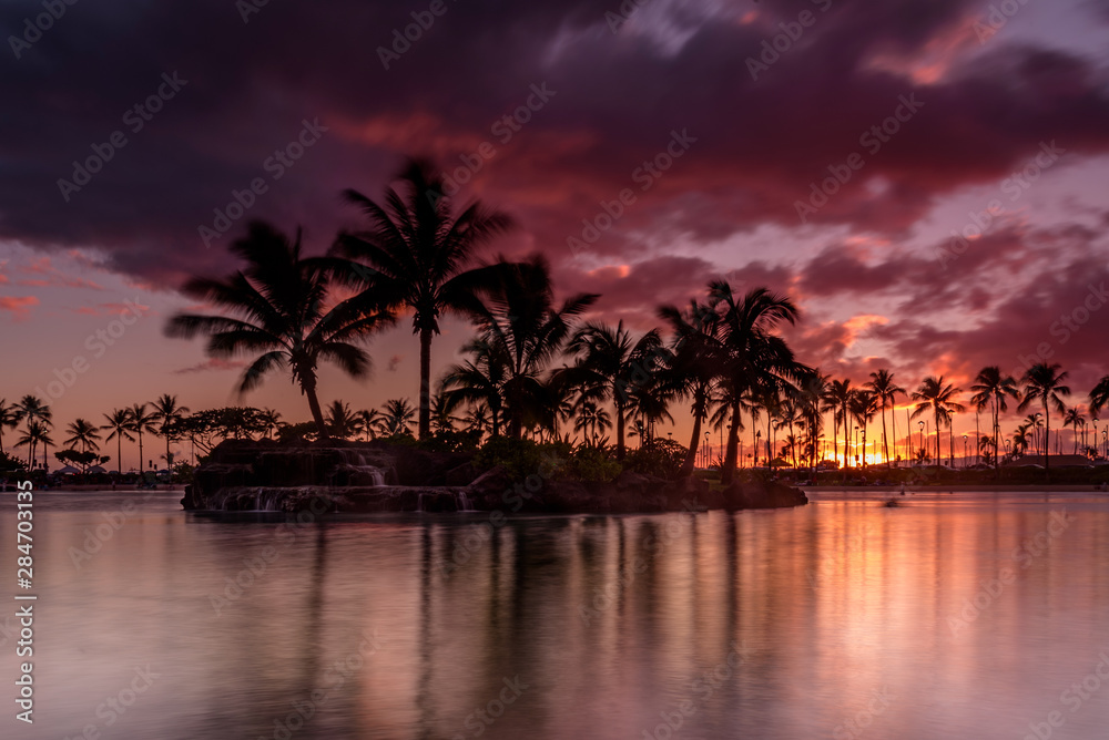 Obraz premium Sun setting on an Hawaiian beach. The sky is purple, red and orange, as the sun dips behind the silhouetted palm trees, on Oahu, Hawaii