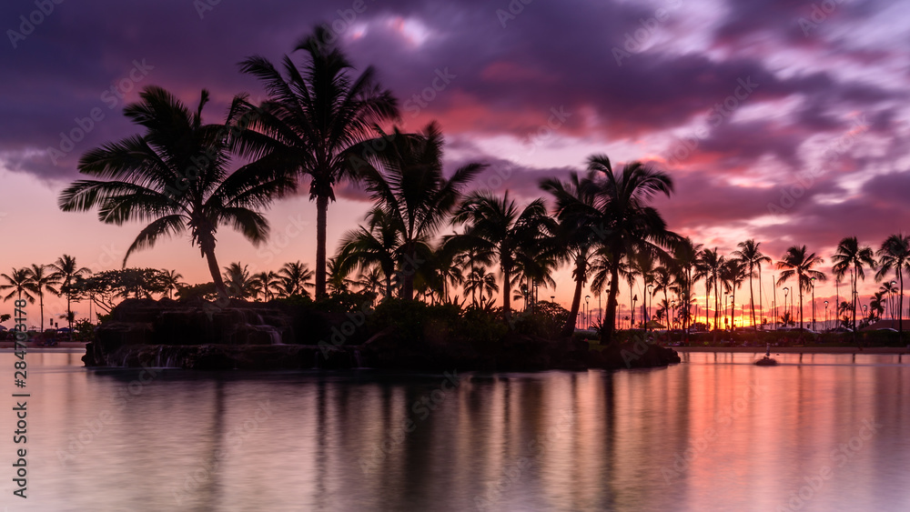Sun setting on an Hawaiian beach. The sky is purple, red and orange, as the sun dips behind the silhouetted palm trees, on Oahu, Hawaii 