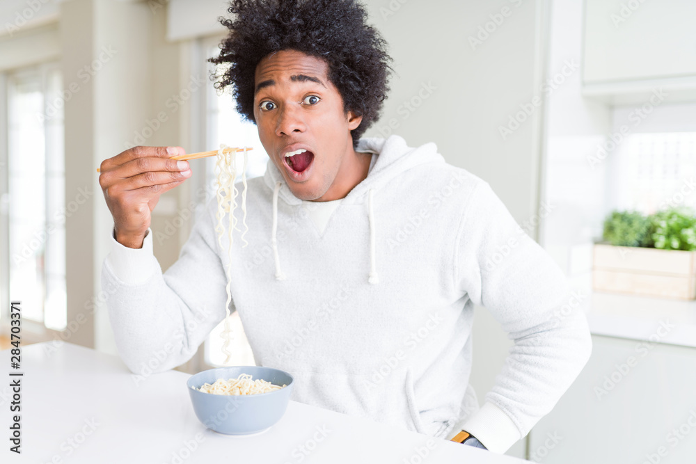 African American man eating asian noodles using chopsticks at home scared in shock with a surprise face, afraid and excited with fear expression African American man eating asian noodles using chopsticks at home scared in shock with a surprise face, afraid and excited with fear expression