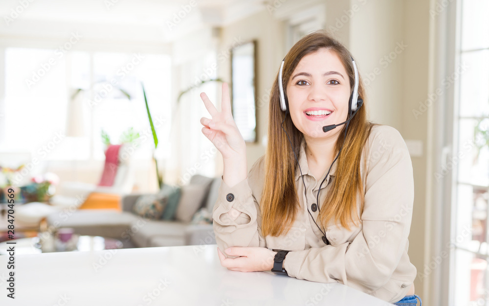 Beautiful young operator woman wearing headset at the office smiling with happy face winking at the camera doing victory sign. Number two. Beautiful young operator woman wearing headset at the office smiling with happy face winking at the camera doing victory sign. Number two.