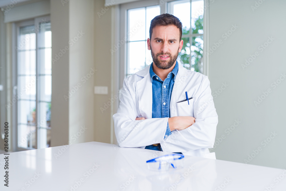 Handsome scientist man wearing white robe and safety glasses skeptic and nervous, disapproving expression on face with crossed arms. Negative person.