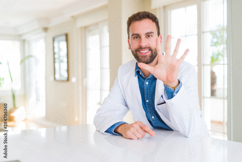 Handsome doctor man wearing medical coat at the clinic showing and pointing up with fingers number five while smiling confident and happy.