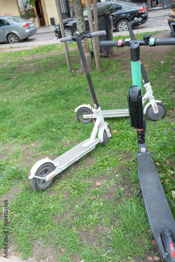 Three electric scooters are standing on the grass in the city center.