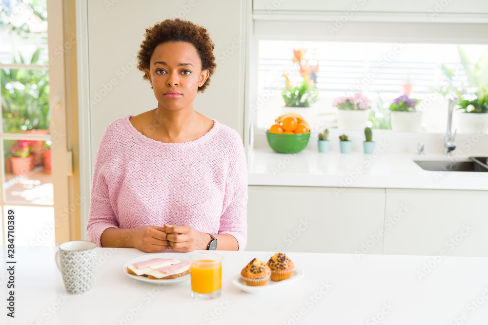 Young african american woman eating breaksfast in the morning at home with serious expression on face. Simple and natural looking at the camera. Young african american woman eating breaksfast in the morning at home with serious expression on face. Simple and natural looking at the camera.