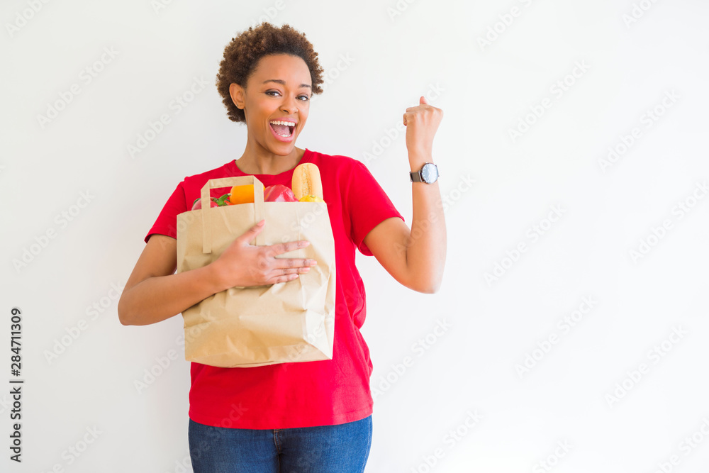 Young african american woman holding paper bag full of fresh groceries screaming proud and celebrating victory and success very excited, cheering emotion Young african american woman holding paper bag full of fresh groceries screaming proud and celebrating victory and success very excited, cheering emotion