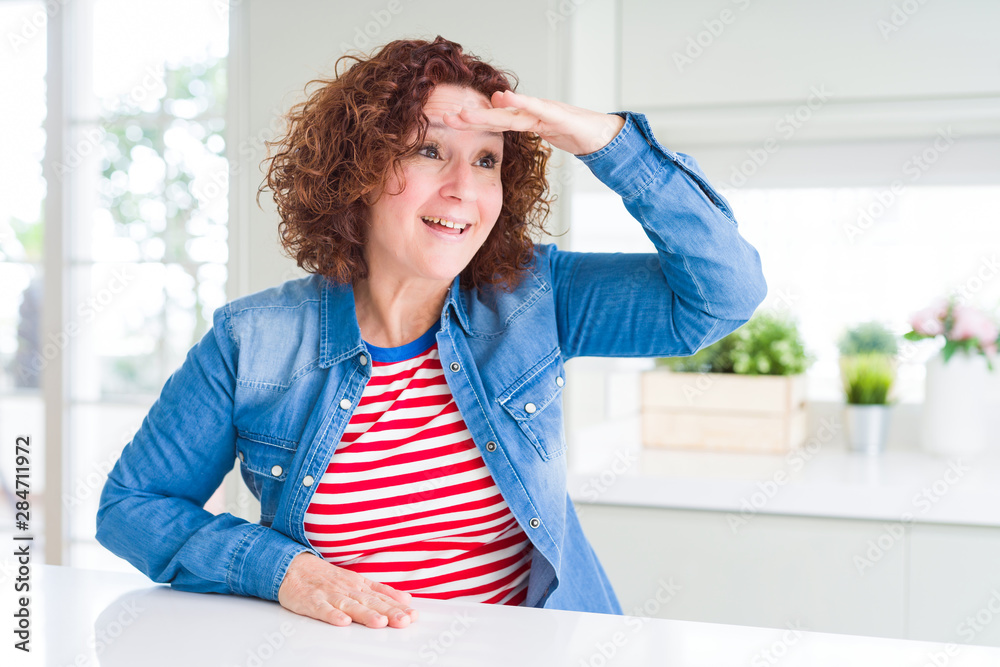 Middle age senior woman with curly hair wearing denim jacket at home very happy and smiling looking far away with hand over head. Searching concept. Middle age senior woman with curly hair wearing denim jacket at home very happy and smiling looking far away with hand over head. Searching concept.